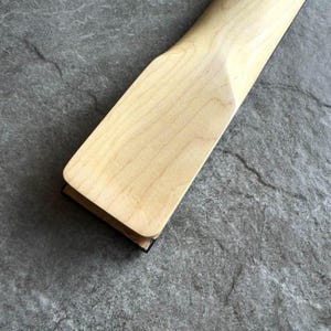 May include: Close-up of a light-colored wooden guitar neck. The neck has a rectangular headstock and a smooth, natural finish. The wood grain is visible, and the neck appears to be new.