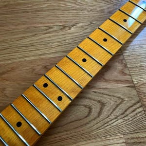 May include: Close-up of a guitar neck with frets and dot inlays. The neck has a light-colored wood finish with a glossy sheen. The frets are silver, and the dot inlays are black. The neck is angled diagonally across a wooden surface.