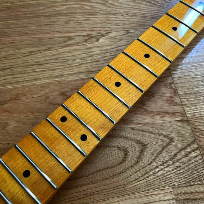 May include: Close-up of a guitar neck with frets and dot inlays. The neck has a light-colored wood finish with a glossy sheen. The frets are silver, and the dot inlays are black. The neck is angled diagonally across a wooden surface.