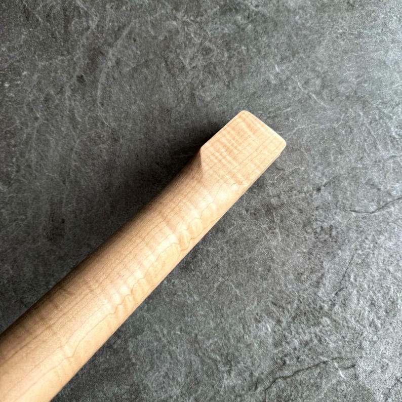 May include: Close-up of a light-colored wooden guitar neck. The wood grain is visible, with a rectangular end. The neck is angled, showing the smooth, finished surface. The background is a textured gray surface.