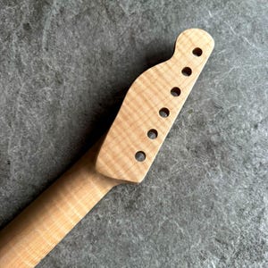 May include: Close-up of a light-colored, unfinished guitar neck. The headstock features six circular holes for tuning pegs. The wood grain is visible, with a natural, unpainted finish. The neck tapers down to a slender profile.