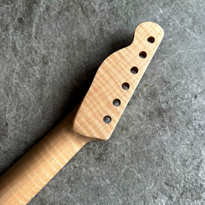 May include: Close-up of a light-colored, unfinished guitar neck. The headstock features six circular holes for tuning pegs. The wood grain is visible, with a natural, unpainted finish. The neck tapers down to a slender profile.