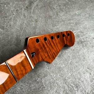 May include: Close-up of a guitar neck and headstock. The headstock is a rich, glossy brown with six tuning peg holes. The neck has a polished finish with frets and a white inlay. The wood grain is visible.