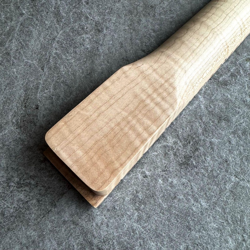 May include: Close-up of a wooden guitar neck, showcasing the natural wood grain and a light, warm color. The neck has a smooth, finished surface and a rectangular shape. The wood appears to be maple, with a slight curl pattern visible.