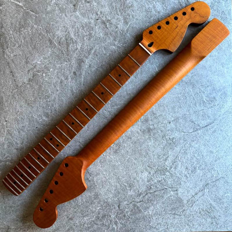 May include: Two unfinished guitar necks, one with frets and tuning peg holes, the other without. Both are made of light brown wood, with a natural finish. The fretboard has silver frets. The necks are positioned diagonally on a gray surface.