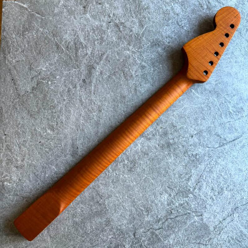 May include: A new, unfinished guitar neck made of light brown wood. The neck has a headstock with six tuning peg holes and a long, slender shaft with a ridged texture. The wood grain is visible throughout the neck.