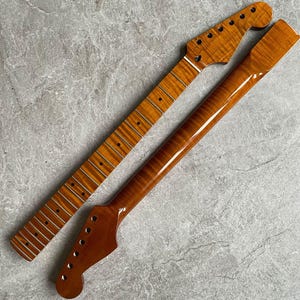 Könnte beinhalten: Zwei unfertige Gitarrenhälse, einer mit Bünden und der andere ohne. Beide bestehen aus hellbraunem Holz mit glänzender Oberfläche. Die Kopfplatten haben Löcher für die Stimmwirbel. Die Holzmaserung ist sichtbar.