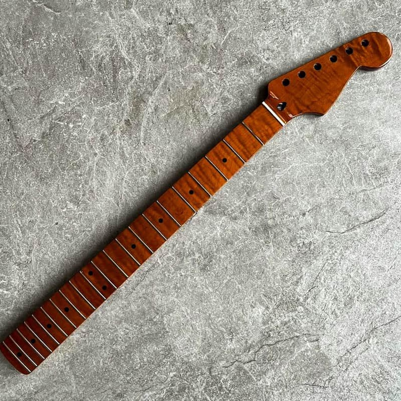 May include: A guitar neck with a flamed maple finish. The neck is a warm brown color with a glossy finish. The fretboard has silver frets and black dot inlays. The headstock has tuning peg holes.