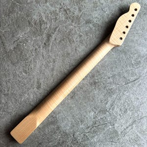 May include: A light-colored, unfinished guitar neck with a headstock featuring six tuning peg holes. The neck is made of wood and has a smooth, natural finish. The neck is angled and ready for installation.