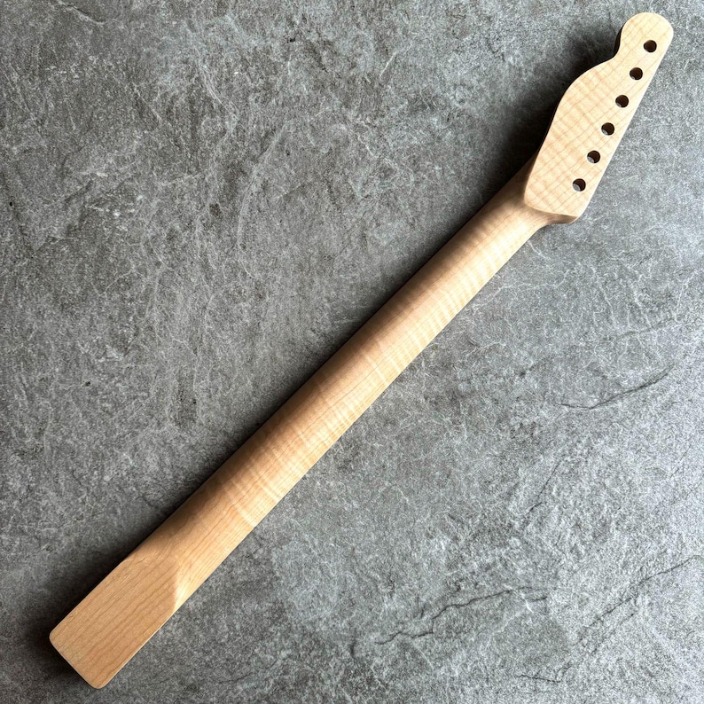May include: A light-colored, unfinished guitar neck with a headstock featuring six tuning peg holes. The neck is made of wood and has a smooth, natural finish. The neck is angled and ready for installation.