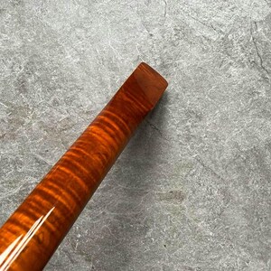 May include: A close-up of a wooden instrument neck with a glossy finish. The wood grain features a flame pattern in shades of orange and brown. The end of the neck is a solid, rectangular shape.