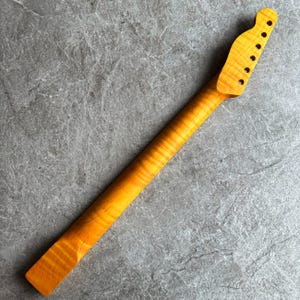 May include: A maple guitar neck with a natural finish. The neck has a headstock with six tuning peg holes and a smooth, flamed grain pattern. The wood has a warm, golden hue.