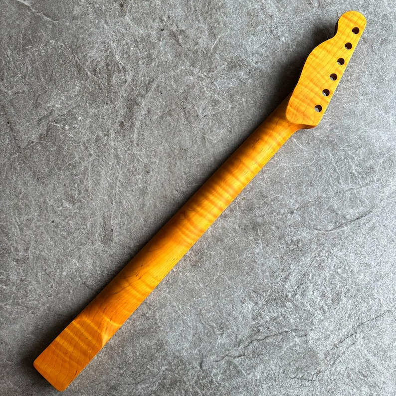 May include: A maple guitar neck with a natural finish. The neck has a headstock with six tuning peg holes and a smooth, flamed grain pattern. The wood has a warm, golden hue.