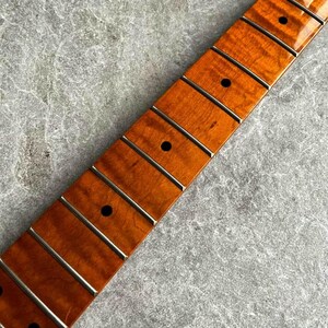 May include: Close-up of a guitar neck with a flamed maple fretboard. The neck has a warm, amber finish with dark fret markers and silver frets. The wood grain is visible, and the frets are evenly spaced.