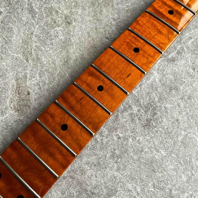 May include: Close-up of a guitar neck with a flamed maple fretboard. The neck has a warm, amber finish with dark fret markers and silver frets. The wood grain is visible, and the frets are evenly spaced.