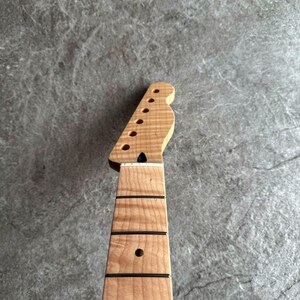 May include: Close-up of a light-colored wooden guitar neck and headstock. The headstock has six tuning peg holes and a unique shape. The neck features black frets and a single dot inlay. The wood grain is visible.