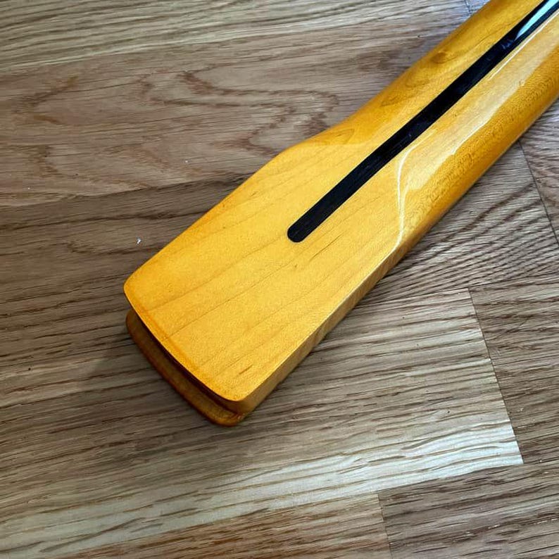 May include: Close-up of a guitar neck with a light yellow finish. A black stripe runs down the center of the neck. The wood grain is visible. The neck is angled towards the bottom left.