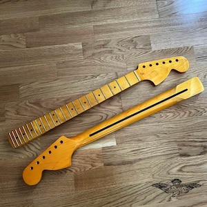 May include: Two unfinished guitar necks, one with frets and tuning peg holes, the other with a black stripe. Both are light-colored wood, lying on a wooden surface. The necks are for a stringed instrument.