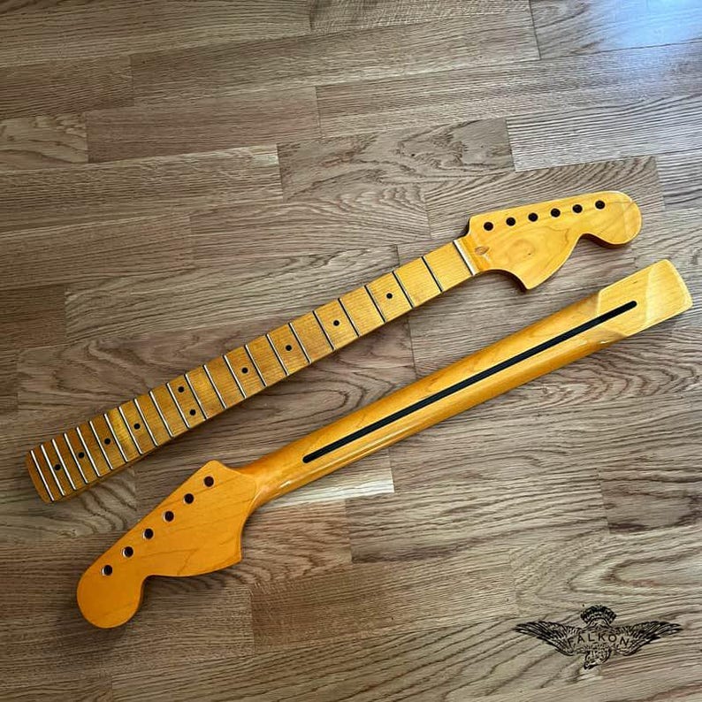 May include: Two unfinished guitar necks, one with frets and tuning peg holes, the other with a black stripe. Both are light-colored wood, lying on a wooden surface. The necks are for a stringed instrument.