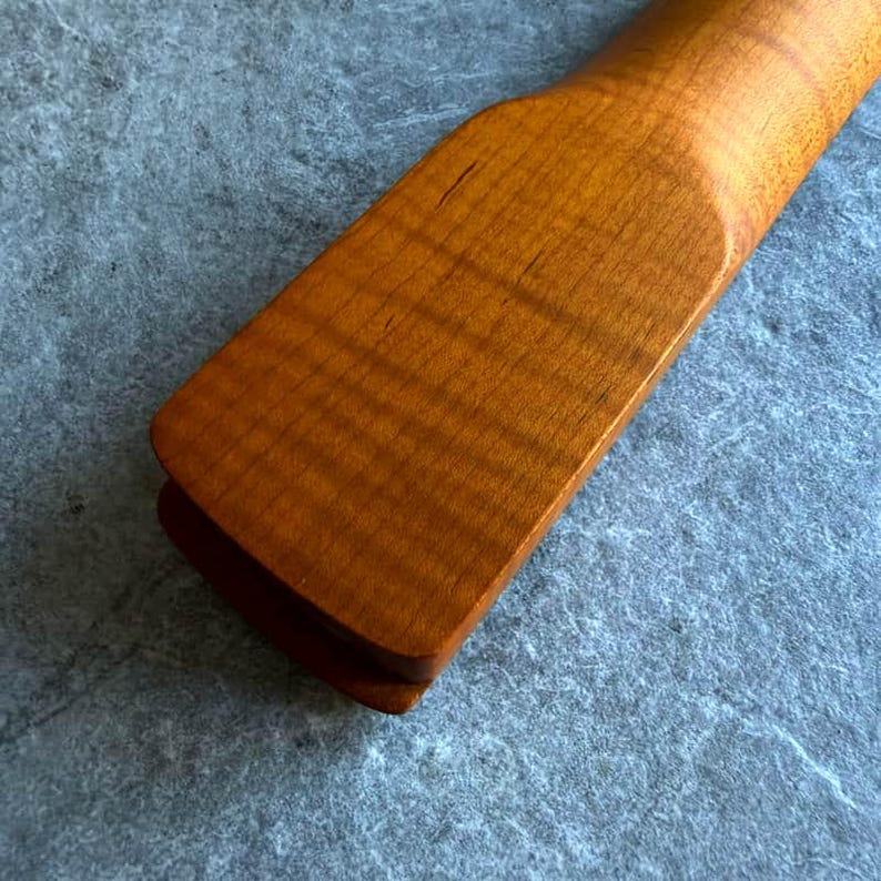 May include: Close-up of a wooden guitar neck, showcasing the natural wood grain and a warm, amber finish. The neck has a smooth, rounded profile and a rectangular end. The wood's texture is visible, with subtle variations in color.