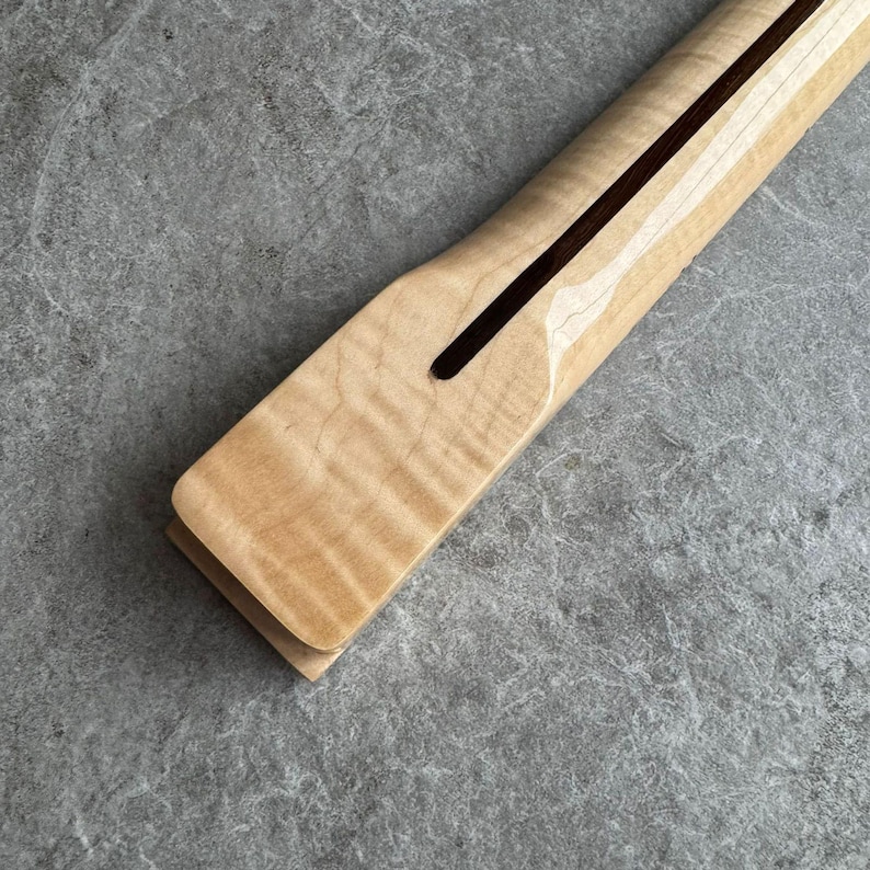 May include: Close-up of a maple guitar neck with a dark brown fretboard slot. The wood grain is visible, and the neck has a smooth, polished finish. The neck is angled, showing the headstock end.