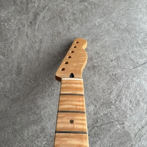 May include: A light-colored, unfinished guitar neck with a headstock featuring tuning peg holes. The fretboard has dark lines and a single dot inlay. The wood grain is visible, and the neck is set against a gray background.