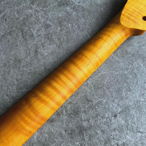 May include: A close-up of a guitar neck made of figured maple. The wood grain has a prominent flame pattern with alternating bands of light and dark amber hues. The neck has a smooth, polished finish and a single tuning peg hole.
