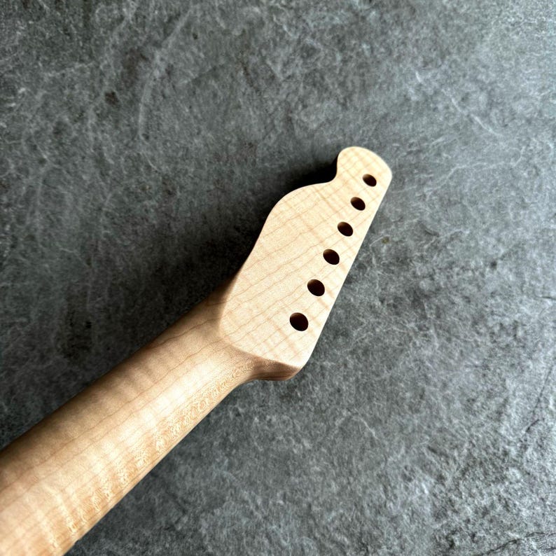 May include: A light-colored wooden guitar neck with a headstock featuring six tuning peg holes. The neck has a smooth, natural finish and a slightly curved shape. The wood grain is visible, adding to the instrument's aesthetic.