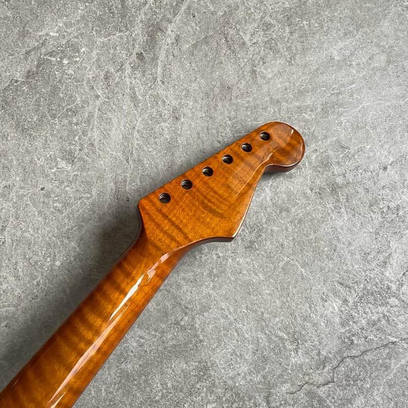 K&ouml;nnte beinhalten: Nahaufnahme eines Gitarrenhalses mit gl&auml;nzendem, bernsteinfarbenem Finish. Die Kopfplatte hat sechs L&ouml;cher f&uuml;r die Stimmwirbel. Die Holzmaserung erzeugt ein gestreiftes Muster. Der Hals ist abgewinkelt und zeigt seine volle L&auml;nge.