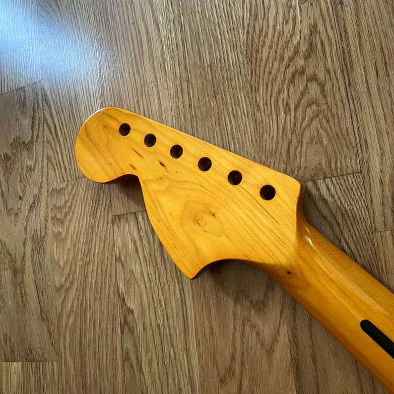 May include: A light-colored wooden guitar neck with six tuning peg holes. The neck has a smooth finish and a dark stripe on the back. The headstock has a classic shape, and the wood grain is visible.