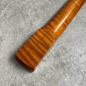 K&ouml;nnte beinhalten: Nahaufnahme eines h&ouml;lzernen Gitarrenhalses mit gl&auml;nzendem, bernsteinfarbenem Finish. Die Holzmaserung ist sichtbar und erzeugt ein gestreiftes Muster. Der Hals ist abgewinkelt und zeigt die Kopfplatte und die glatte, polierte Oberfl&auml;che.