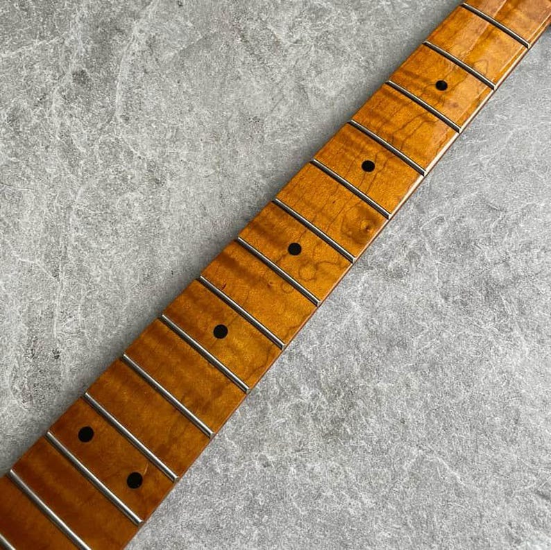 K&ouml;nnte beinhalten: Nahaufnahme eines Gitarrenhalses mit geflammter Ahorn-Oberfl&auml;che. Er hat silberne B&uuml;nde und schwarze Punkteinlagen. Die Holzmaserung ist sichtbar und zeigt eine warme, bernsteinfarbene Farbe.