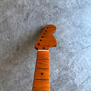 May include: A guitar neck with a natural wood finish. The headstock features tuning peg holes and a curved shape. The fretboard has dark dots and lines. The wood grain is visible throughout the neck.