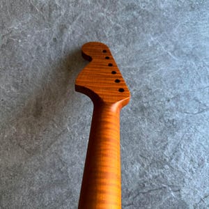 May include: Close-up of a guitar neck and headstock. The wood has a warm, amber tone with visible grain patterns. The headstock features six tuning peg holes. The neck tapers down, showcasing the wood's natural texture and color.