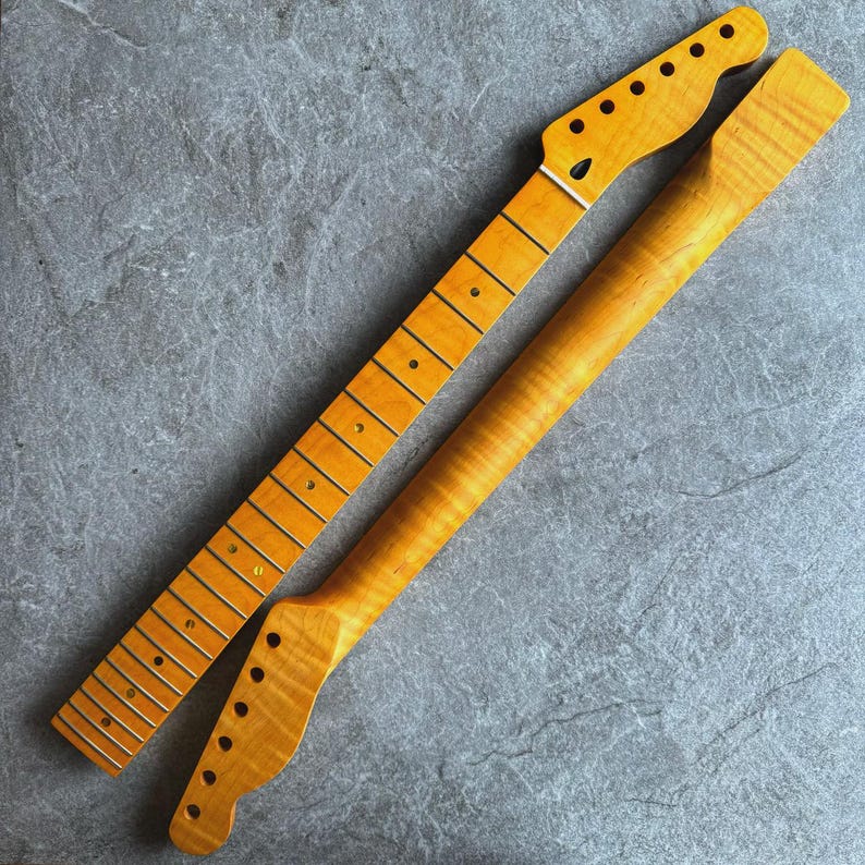 May include: Two unfinished guitar necks, one with frets and dot inlays, the other without. Both are made of light-colored wood with a natural finish. The headstocks have tuning peg holes. The necks are angled and lie on a gray surface.