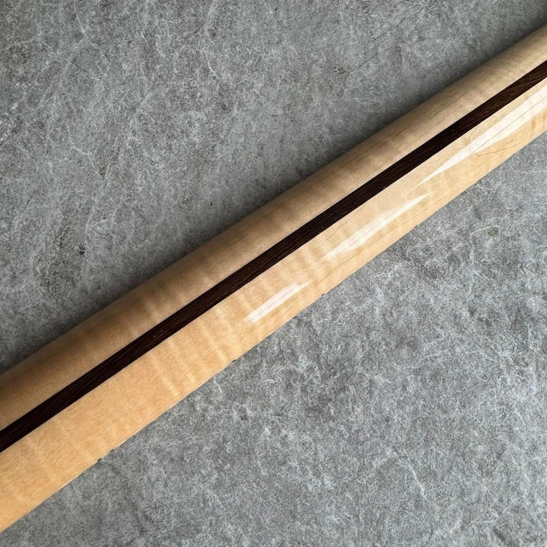 May include: Close-up of a guitar neck with a light-colored wood grain and a dark brown stripe down the center. The neck has a glossy finish, reflecting light. The wood grain has a wavy pattern.