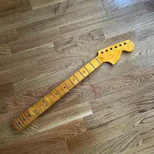 May include: A guitar neck with a natural wood finish. The neck has frets, dot inlays, and tuning peg holes. The wood grain is visible, and the neck is angled across a wooden surface.