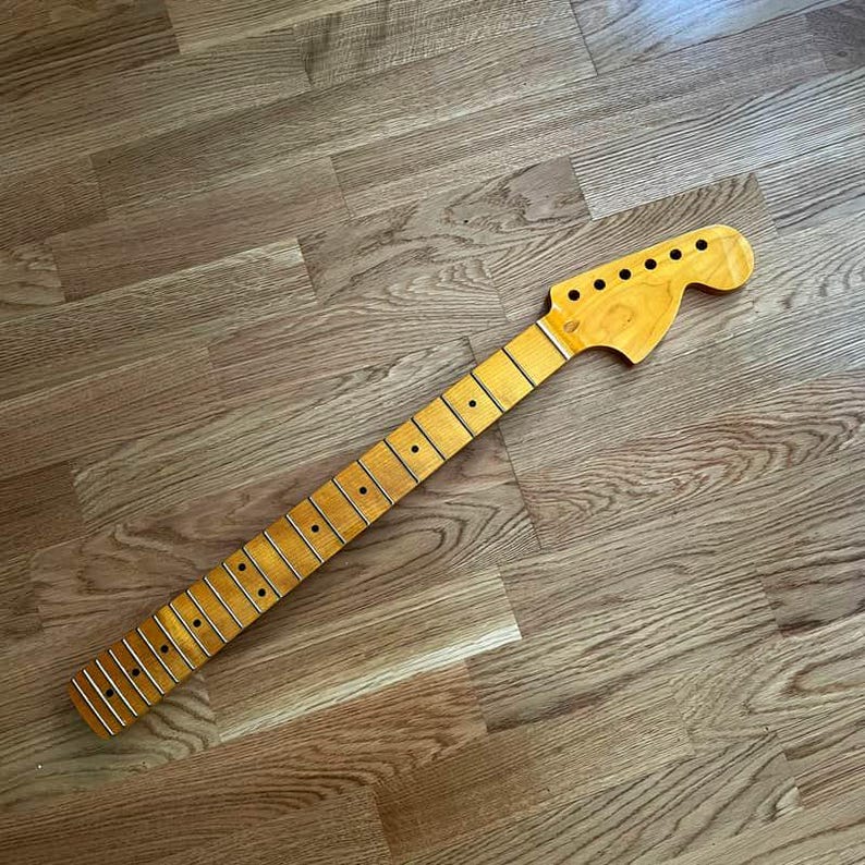 May include: A guitar neck with a natural wood finish. The neck has frets, dot inlays, and tuning peg holes. The wood grain is visible, and the neck is angled across a wooden surface.