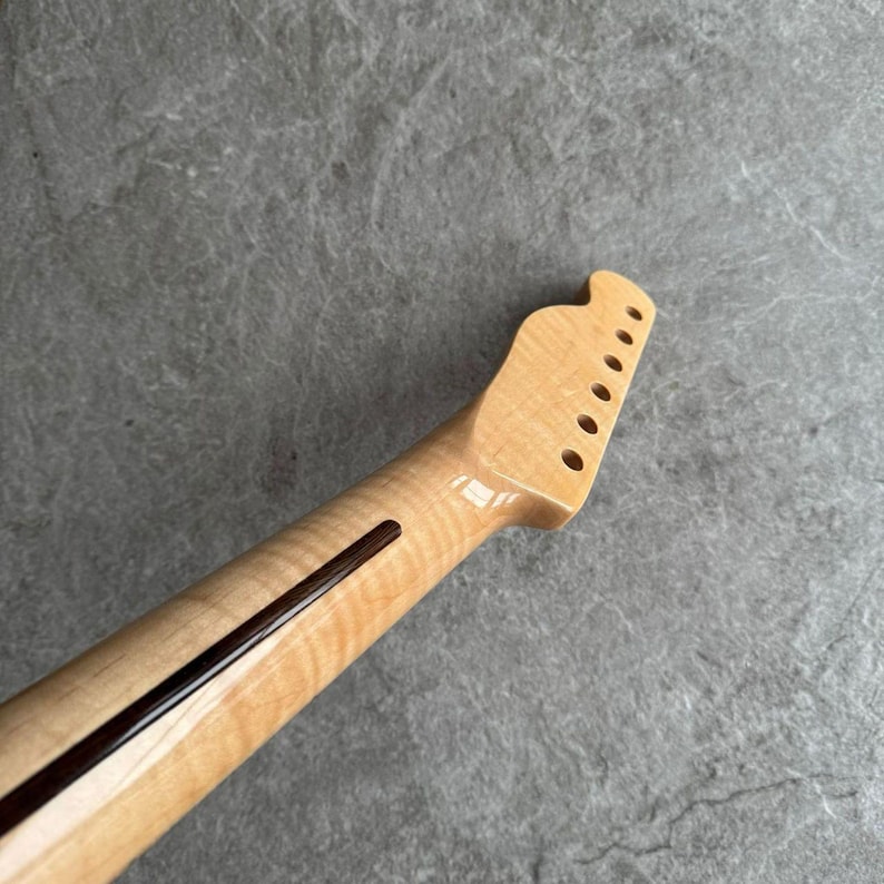 May include: A guitar neck made of light-colored wood, featuring a dark stripe down the center and six tuning peg holes. The headstock has a rounded shape. The wood grain is visible.