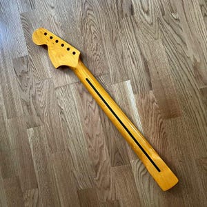 May include: A new, unfinished guitar neck made of light-colored wood. The neck has a headstock with six tuning peg holes and a long, slender neck with a black stripe down the center. The wood grain is visible.