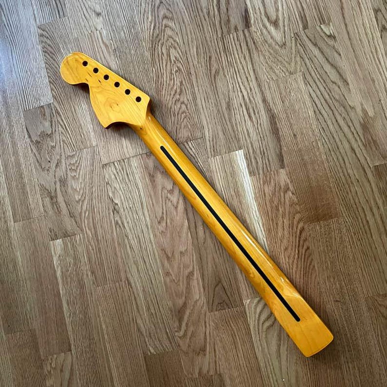 May include: A new, unfinished guitar neck made of light-colored wood. The neck has a headstock with six tuning peg holes and a long, slender neck with a black stripe down the center. The wood grain is visible.