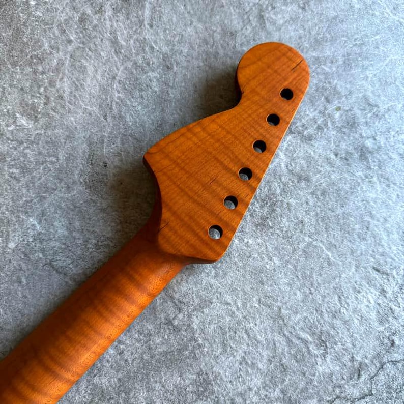 May include: A close-up of a guitar neck and headstock made of light brown wood. The headstock has six circular holes for tuning pegs. The wood grain is visible, and the finish is smooth and polished.