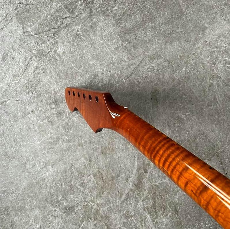 May include: A close-up of a guitar neck, showcasing a rich, reddish-brown wood grain. The neck features tuning peg holes and a smooth, varnished finish. The wood grain has a distinctive, wavy pattern.