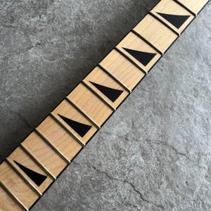 May include: Close-up of a guitar neck with light wood frets and black triangle inlays. The frets are separated by thin metal strips. The neck is set against a gray, textured background.
