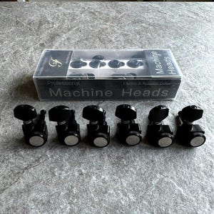 May include: Six black metal guitar machine heads arranged in a row in front of a clear plastic box. The box features black labels with white text reading Professional Machine Heads for Electric and Acoustic Guitar and a gold F logo.