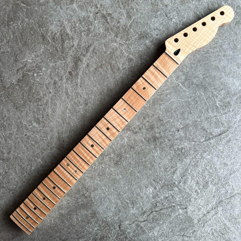 May include: A maple wood guitar neck with a natural finish. The neck features a headstock with pre-drilled holes for tuning pegs and a fretboard with inlaid fret markers. The wood grain is visible.