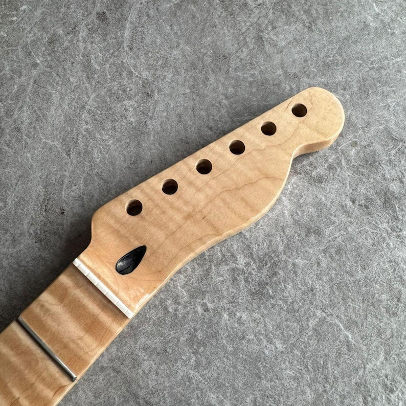 May include: A light-colored, unfinished guitar neck with six tuning peg holes and a fretboard. The wood grain is visible, and the neck is ready for assembly. The headstock has a classic shape.