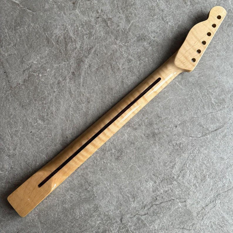 May include: A light-colored, unfinished guitar neck with a headstock featuring six tuning peg holes. The neck has a dark stripe down the center and is made of wood. The wood grain is visible.