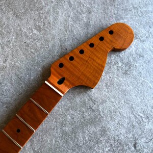 May include: A close-up of a guitar neck and headstock. The headstock is a light brown color with six black tuning peg holes. The neck is a darker brown with silver frets. The wood grain is visible.