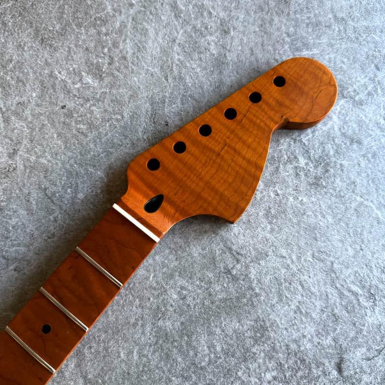 May include: A close-up of a guitar neck and headstock. The headstock is a light brown color with six black tuning peg holes. The neck is a darker brown with silver frets. The wood grain is visible.